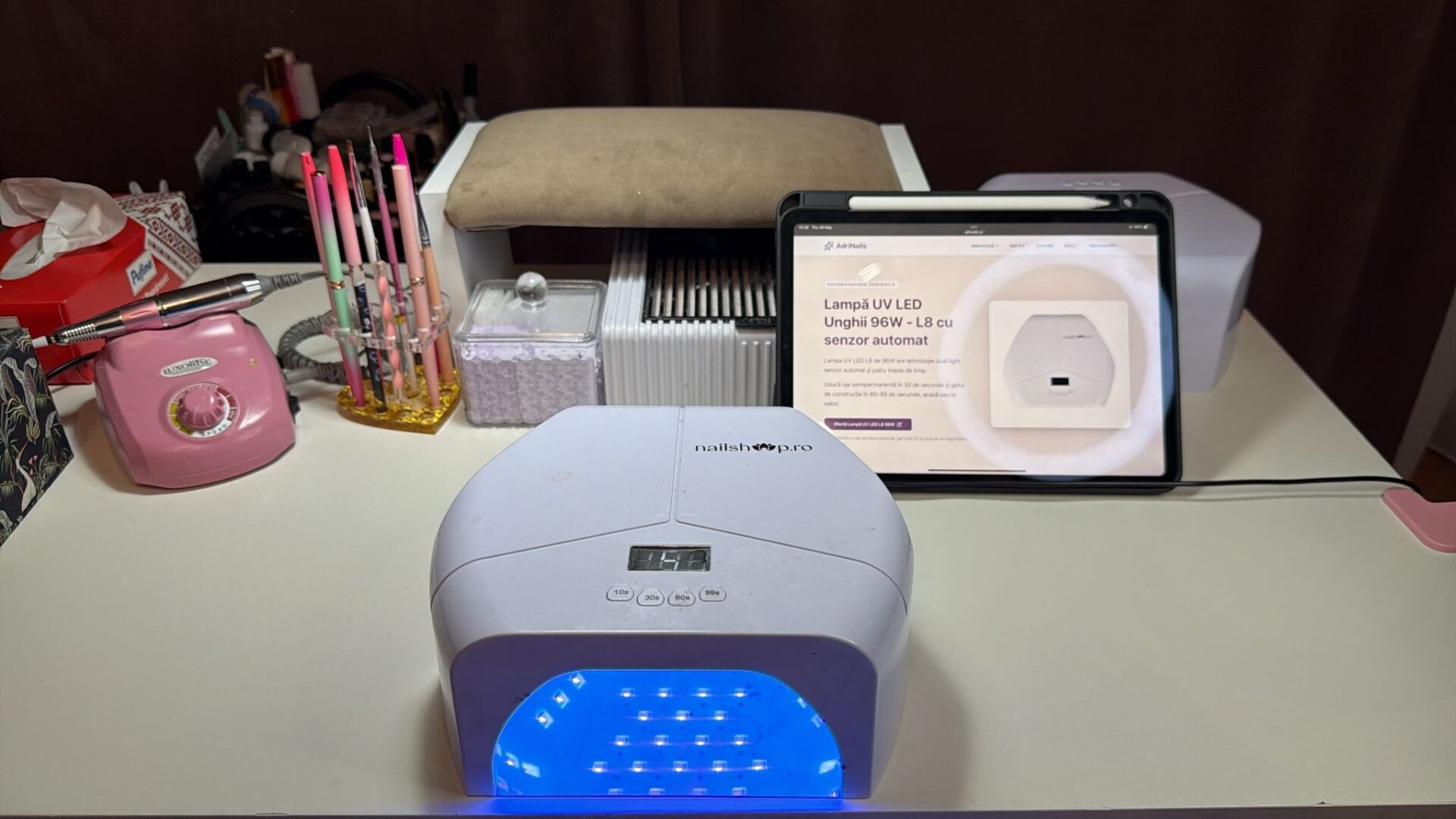 Lampa UV LED L8 96W aprinsă pe masa de manichiură, lângă freză și tabletă cu pagina AdriNails